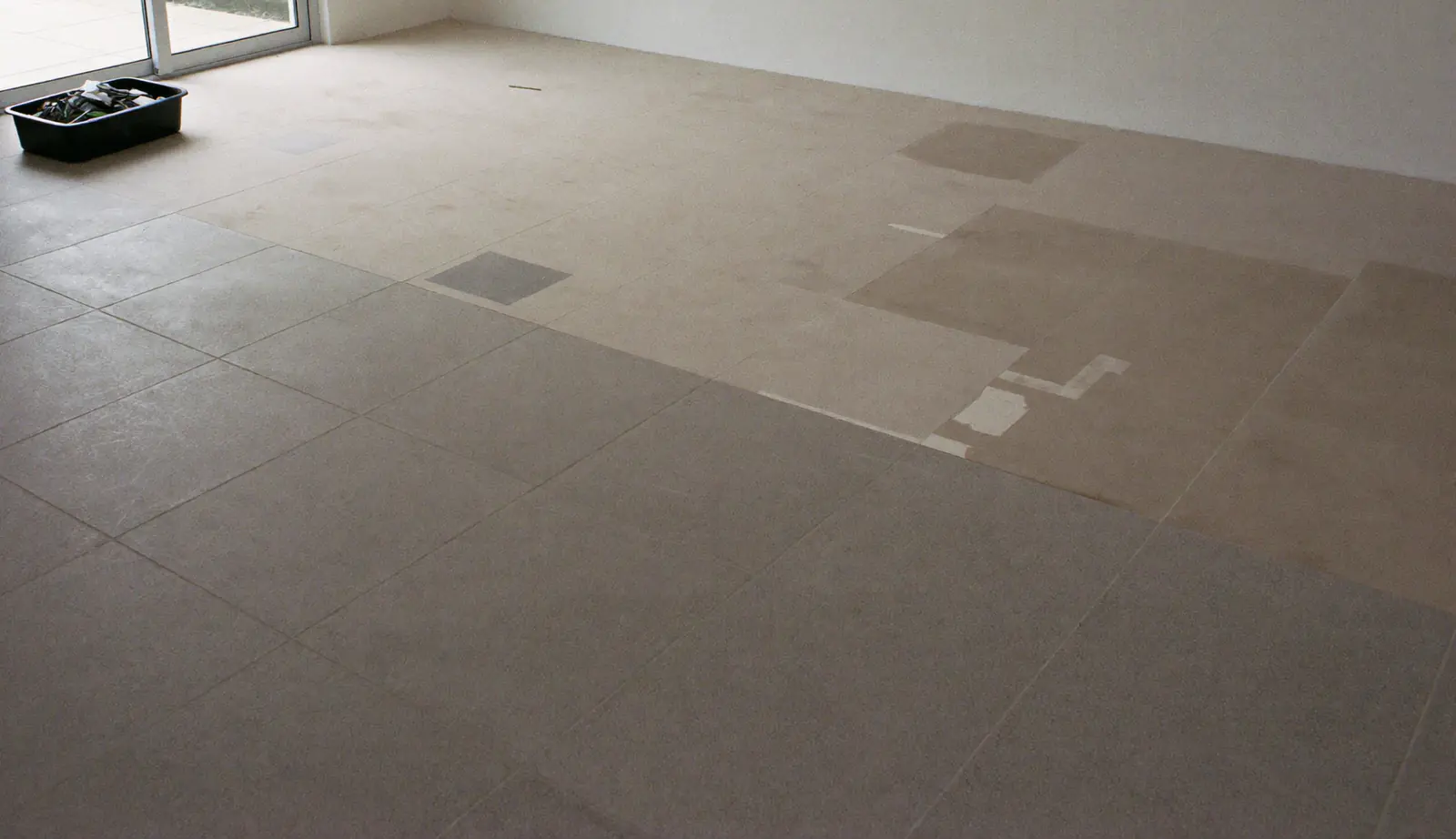Lege woonkamer in een rijwoning uit de jaren negentig waar een deel van de grijze tegelvloer al is verwijderd en de lichte dekvloer zichtbaar is