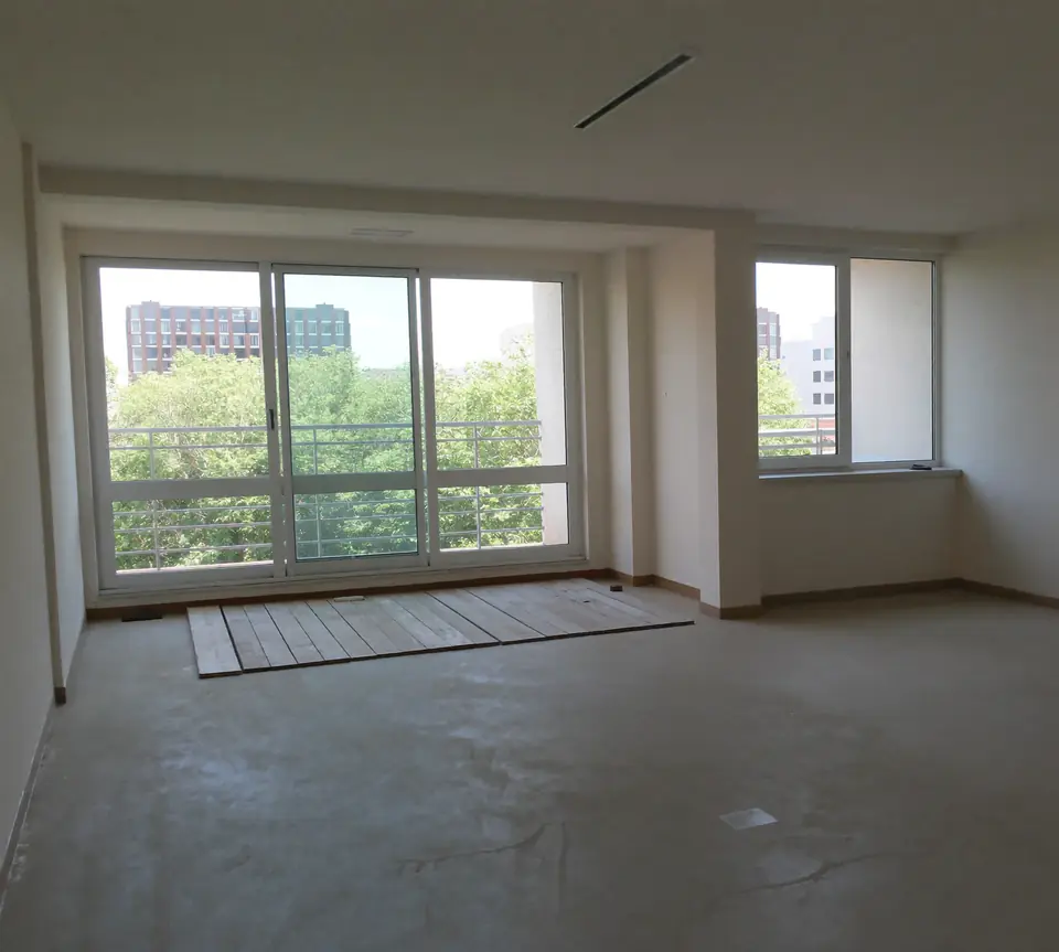 Lege woonkamer in een appartement met een kale betonvloer en grote ramen naar een balkon met uitzicht op andere flats