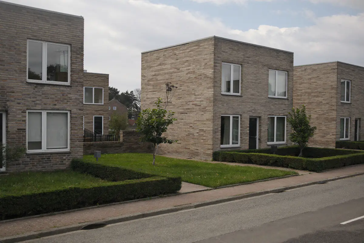 Straat met moderne bakstenen woningen met platte daken, kleine voortuinen met hagen en jonge bomen in een nieuwbouwwijk