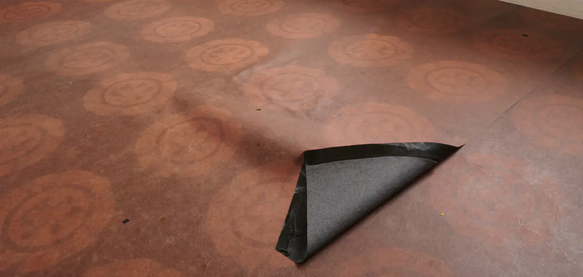 Oud bruin vloerzeil met een rond motief waarvan een hoek is opgekruld en de grijze viltachtige onderlaag zichtbaar is